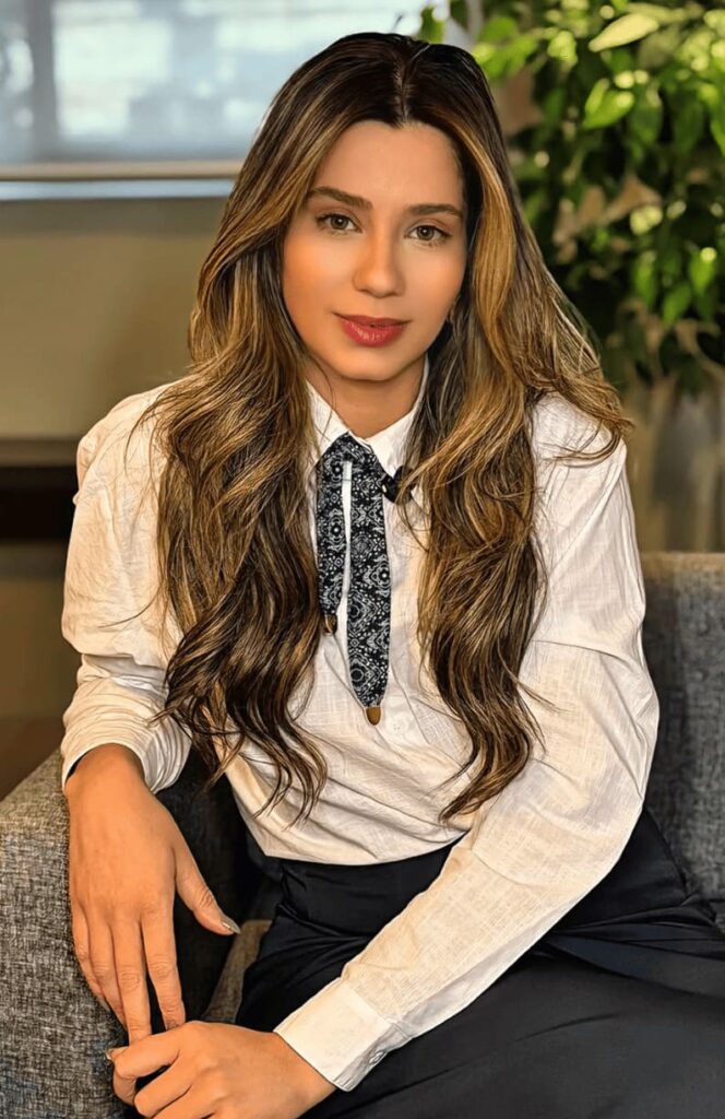 Mulher de cabelos longos com luzes, vestindo camisa social branca e gravata feminina estilizada, sentada em um sofá.